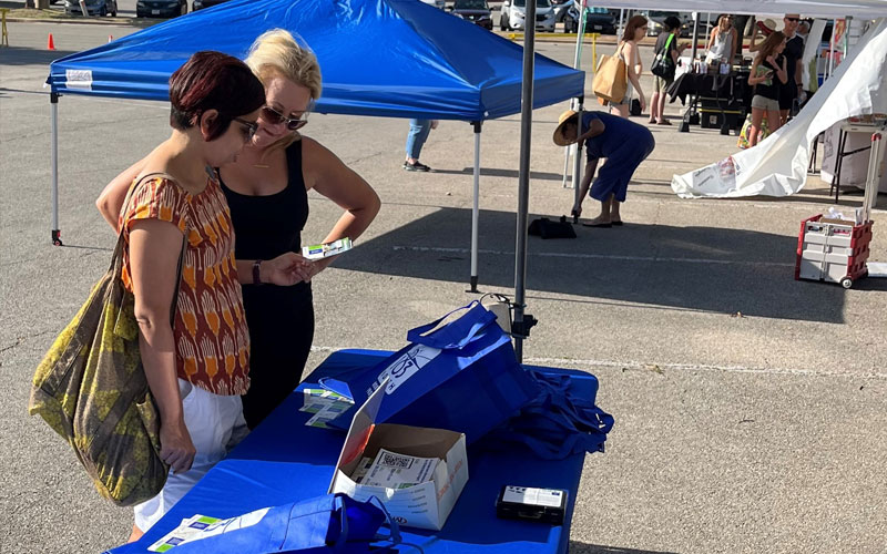 Two women stand together looking at a table of items at an outdoor market. 