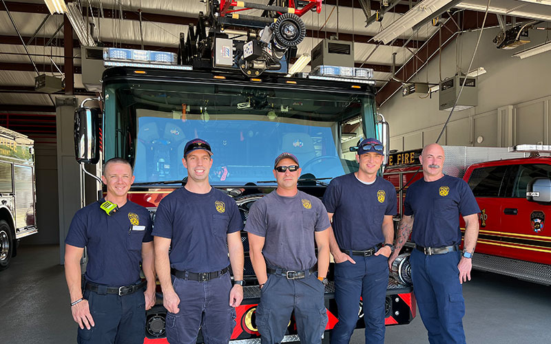 Five firemen in similar blue t-shirts stand in front of a fire truck at the fire house. 