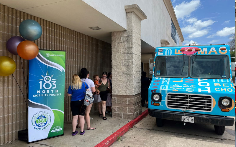 A blue ice cream truck is parked next to a beige building with people standing under the building's overhang. 