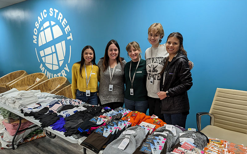 Five women stand together behind a table of items.