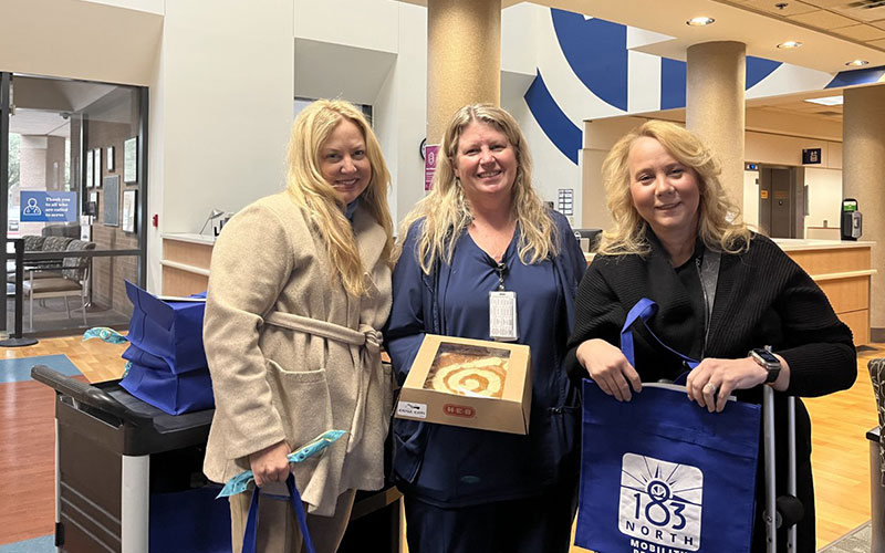 Three women pose for a photo with one holding a boxed pie and another holding a blue bag. 