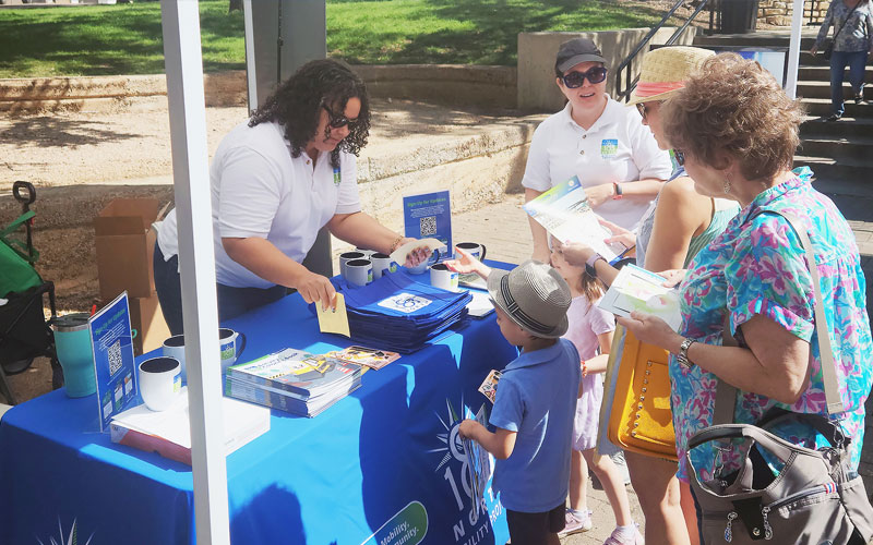 A group of people including a child and adults stand around a table with a woman handing out information.