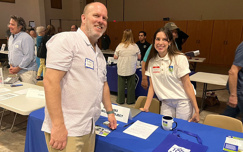 A man and women pose for a photo while standing on either side of a table. There are people mingling in the background. 