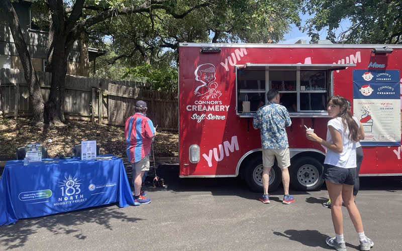 A man orders from a red ice cream truck in a parking lot. Two people are standing nearby. 