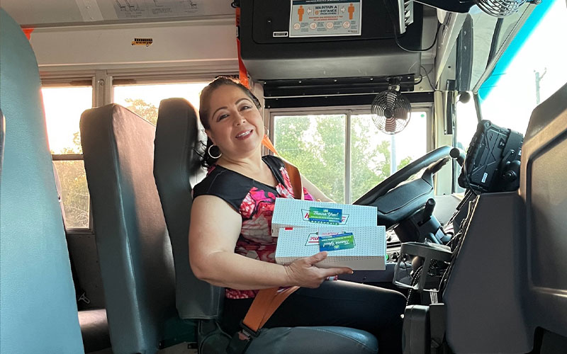 A women holds two boxes of donuts while sitting in the driver seat of a school bus. 