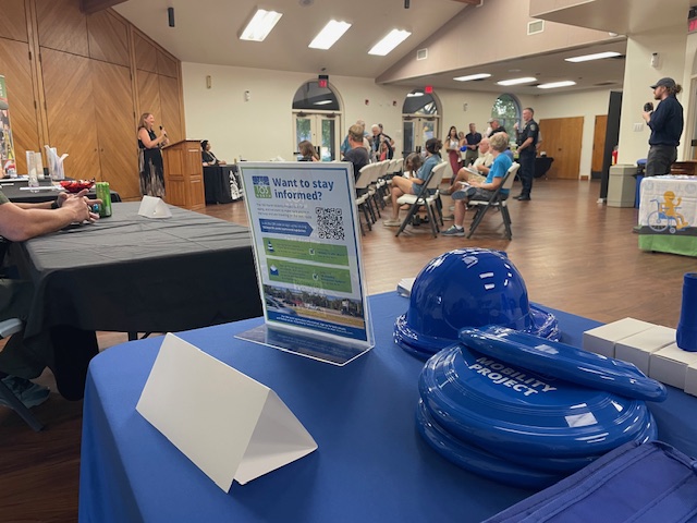 Audience listens to womn apeaking at podium, photo taken from angle of informational table showing 183 North Mobility Project materials in the foreground