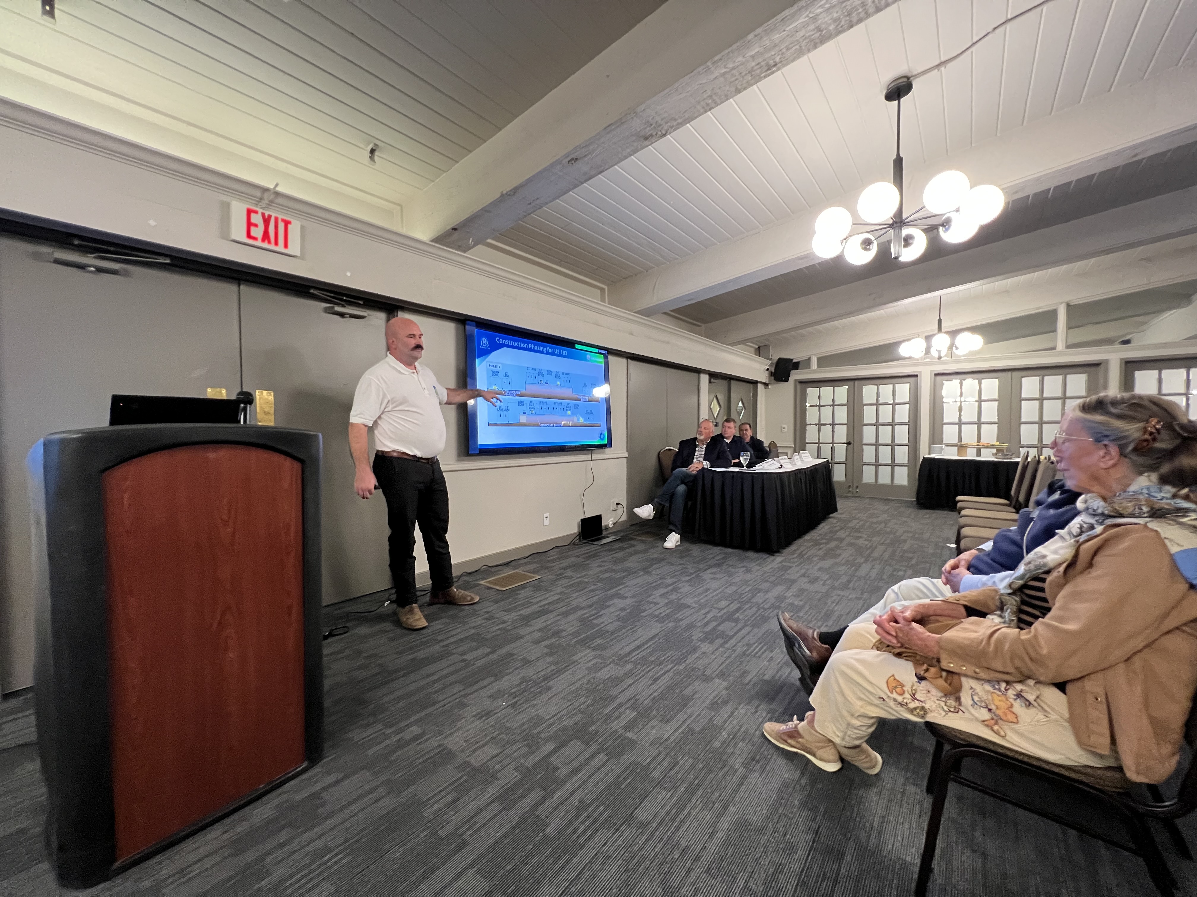 A man gives a presentation standing at the front of a room. Two people in the front row are watching him. 